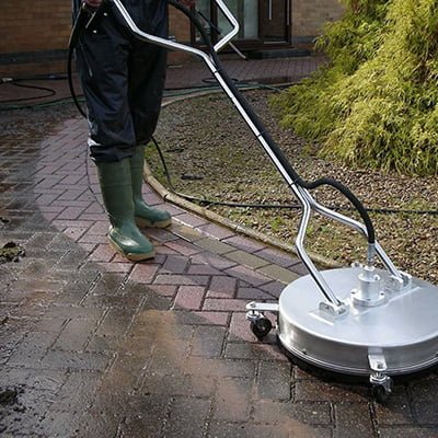 A person in rubber boots uses a high-pressure washer with a surface attachment to clean a dirty brick pavement, showing a clear before and after effect.